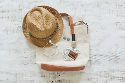 A calm overhead flat lay of warm-weather travel essentials: a woven straw hat, cream canvas tote bag with brown leather trim, small jewellery, nail polish and a charging cable on white wood.